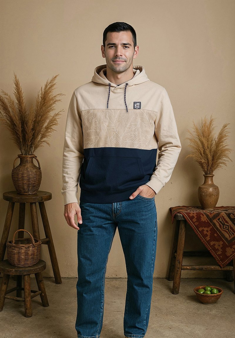 Hoodie met beige bovenkant, marineblauwe onderkant en een bladpatroon; gecombineerd met een blauwe spijkerbroek. Model staat met handen in de zakken, neutrale achtergrond.