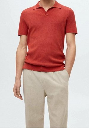 Mężczyzna w dopasowanej, czerwonej koszulce polo z krótkim rękawem, wsadzonej do beżowych, plisowanych spodni, z ręką częściowo w kieszeni.