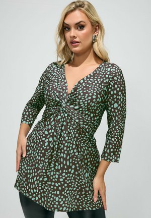 Blonde vrouw die een bruine tuniek draagt met lichtblauwe onregelmatige polka dots, voorzien van een gedraaide knoop en driekwartmouwen.