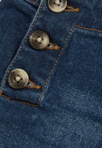 Középkék árnyalatú denim szövet, három kör alakú, áttetsző gombbal és kontrasztos narancssárga varrással az varráson.