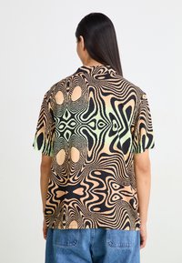 Chemise à manches courtes en noir, crème et vert clair avec un motif abstrait en spirale. Le tissu a une texture lisse et une coupe décontractée.