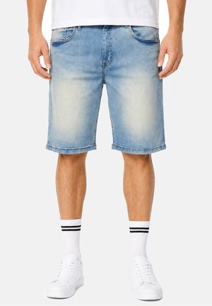 Mann trägt hellblaue Jeansshorts, weiße Sneakers und weiße Socken mit zwei schwarzen Streifen, steht vor einem einfarbigen Hintergrund.