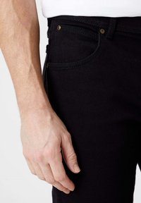 Jeans in denim nero con una vista ravvicinata della zona della tasca laterale e della mano. Presentano accessori in oro e dettagli di cucitura visibili.