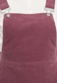 Peto de pana en color rosa empolvado con estrías verticales, que presenta hebillas de metal y una textura suave y lisa.