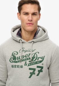 Grå hættetrøje med grøn grafisk tekst, der læser "SuperDry" og "Boot Co" på forsiden. Har snører og et blødt, tekstureret stof.