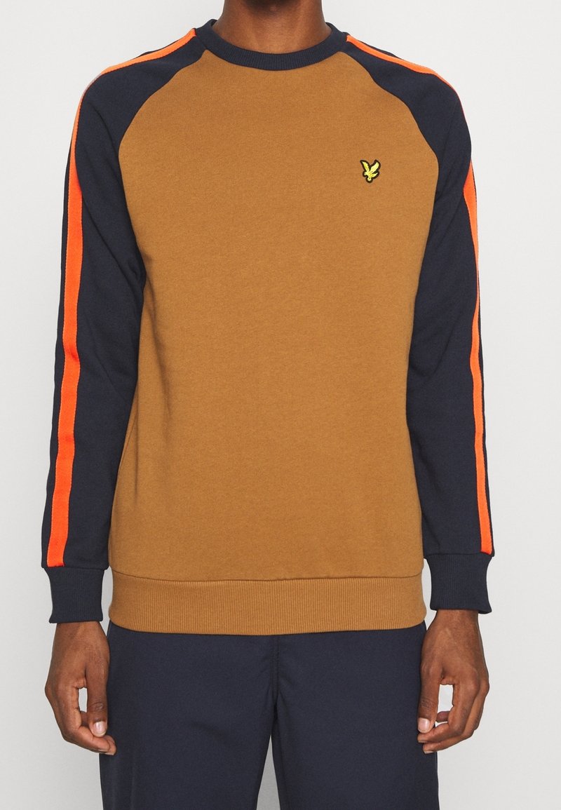 Homme portant un sweat-shirt marron et marine avec des bandes orange sur les manches et un petit logo d'oiseau jaune sur la poitrine gauche.