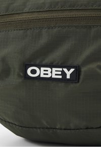 Olívazöld szövet hátizsák, texturált felülettel; felső cipzárral és egy téglalap alakú fekete logóval, amelyen a "OBEY" felirat áll fehér színnel.