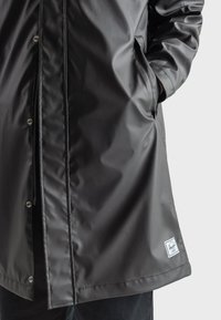 Black waterproof jacket with a smooth finish, snap buttons, side pockets, and a branded label at the hem. Lightweight and functional design.