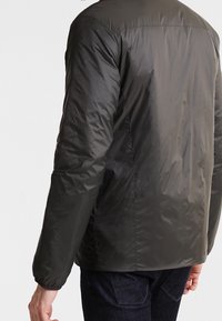 Chaqueta aislante de color verde oscuro con una textura suave y brillante, que presenta un diseño simple y puños elásticos. Costuras visibles a lo largo de las juntas.