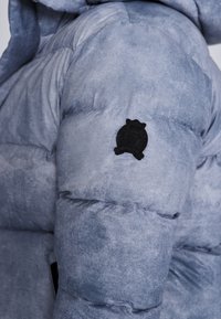 Polstret blå-grå jakke med en tekstureret overflade, udstyret med et rundt sort logo-patch på ærmet og en hætte.