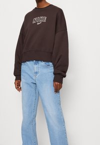 Osoba w ciemno-brązowej bluzie Nike z logo i jasnoniebieskich spodniach z wysokim stanem, stojąca na tle jednolitej białej ściany.