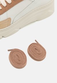 Vue partielle d'une chaussure de sport blanche et beige au-dessus de deux lacets beiges enroulés sur un fond blanc.