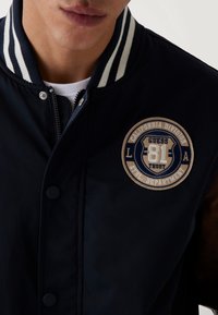 Chaqueta bomber navy con cuello acanalado blanco a rayas, cremallera frontal y un parche circular beis con texto azul en el pecho.