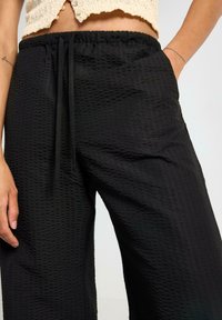 Pantaloni neri in tessuto strutturato con vita elastica e lacci. Presentano tasche laterali e un design ampio con gambe larghe.