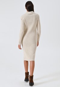Beige, geribbelde gebreide midi-jurk met een rolkraag en lange mouwen, aansluitende silhouet, gecombineerd met bruine enkelboots.