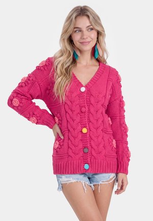 Kvinde med langt blondt hår iført en lys pink chunky strikket cardigan med farverige knapper og lyseblå slidte denimshorts.