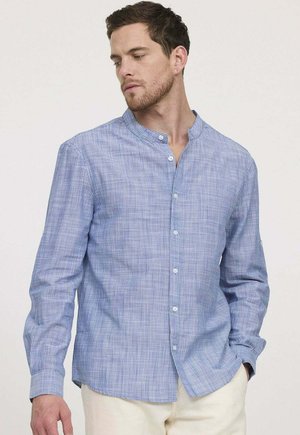 Homme portant une chemise à manches longues à carreaux bleu clair avec col mandarin et pantalon crème, regardant légèrement vers le bas sur sa gauche.