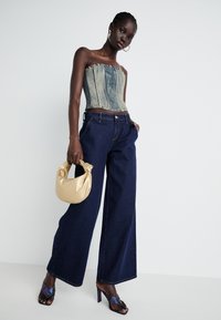 Denim top bez ramínek, široké tmavě modré džíny a krémová saténová kabelka. Modelka má na sobě iridiscentní sandály na podpatku; hladké textury.