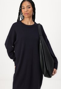 Pulôver oversized azul-marinho com gola redonda e ombros caídos, combinado com uma grande mala de ombro preta em couro. Textura suave e ajuste relaxado.