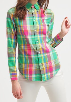 Vrouw draagt een blouse met lange mouwen en knopen met een groen, roze, geel en grijs geruit patroon, gecombineerd met een witte broek.