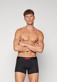 Sorte boksershorts laget av elastisk stoff, med en rød livline med "PUMA"-logo. Designet er tettsittende og har en midt-lårskutt.