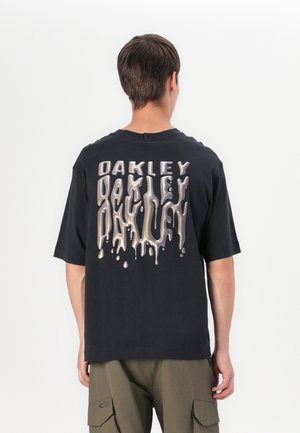 Jeune homme portant un t-shirt noir avec un texte métallique dégoulinant "Oakley" dans le dos et un pantalon cargo vert olive, debout de dos.