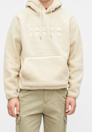 Osoba nosząca beżową bluzę z kapturem z polaru z kieszenią z przodu oraz wytłoczonym napisem „JACK & JONES” na piersi, w połączeniu z jasnymi spodniami typu cargo w kolorze khaki.