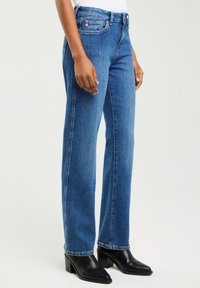 Kobieta ubrana w niebieskie dżinsy o prostych nogawkach i czarne botki na obcasie, stojąca na tle jednolitego tła.