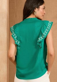Blusa verde senza maniche con spalle arricciate, caratterizzata da ricami bianchi lungo i bordi, texture liscia e vestibilità comoda.