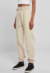 Beige Jogginghose mit einem breiten elastischen Bund, Kordelzugverschluss, seitlichen Taschen und schmal zulaufenden Säumen, kombiniert mit schwarzen High-Top-Sneakern.