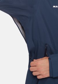 Giacca a vento blu navy in tessuto testurizzato, con cordini regolabili in vita, tasche laterali con zip e polsini elastici per una vestibilità sicura.