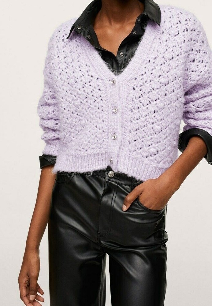 Cardigan en crochet lavande avec boutons en perle porté sur une chemise et un pantalon en fausse cuir noir, une main dans la poche.