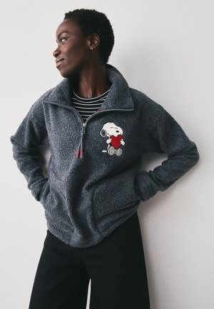 Person iført en grå fleecejakke med Snoopy, der holder et rødt hjerte, står med hænderne på hoften og kigger til venstre mod en ensfarvet baggrund.