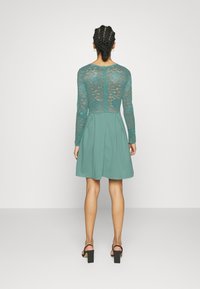 Vestido teal com mangas longas de renda, corpete justo e saia rodada. Apresenta detalhes em renda nas costas e comprimento acima do joelho.