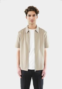 Beige kortermet skjorte med glidelås over en hvit T-skjorte, med en lett avslappet passform og minimalistisk design. Kombinert med svarte bukser.