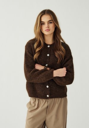 Giovane donna con capelli lunghi e mossi, indossa un cardigan marrone peloso con bottoni e pantaloni beige larghi, braccia incrociate, sfondo neutro.