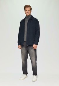 Veste bleu marine foncé avec fermeture à bouton et deux poches avant, portée sur un col roulé gris, associée à un jean gris et des baskets blanches.