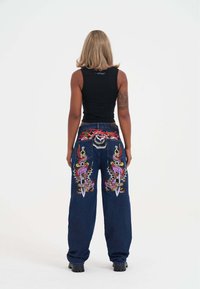 Mørkeblue vidde jeans med livlige broderede designs på baglommerne, herunder sværd og farverige mønstre. Sort tanktop bæres.