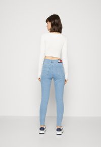 Jeans skinny azul claro de cintura alta, con dos bolsillos traseros y un parche de logo en la cintura, combinados con una blusa blanca de canalé.