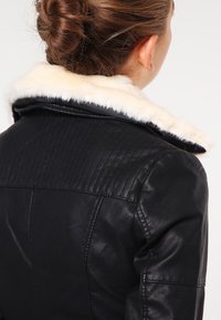 Chaqueta de cuero negra con un cuello de piel sintética blanca, diseño acolchado en los hombros y textura suave. Detalles de costura visibles en la parte posterior.