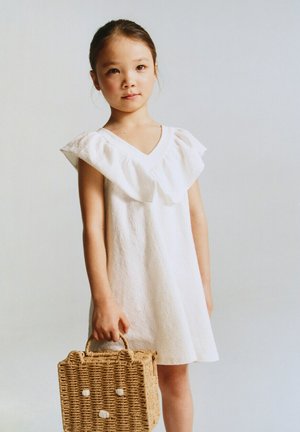 Jeune fille en robe blanche tenant un sac à main en paille tissée avec un motif visage d'ours sur un fond clair uni.