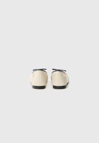 Paire de ballerines beiges avec bouts noirs et petits nœuds noirs, vues de dos sur un fond gris clair.