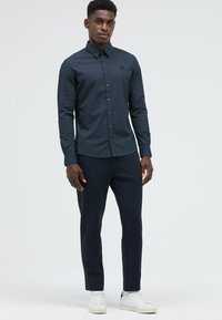 Langärmliges Hemd mit Marine-Muster und Button-Down-Kragen, kombiniert mit dunklen Hosen und weißen Sneakers; strukturierter Stoff und dezentes Logo-Detail.