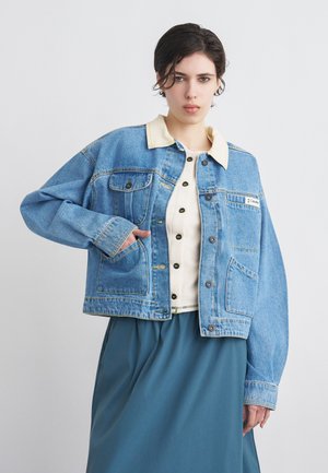 Młoda osoba o krótkich ciemnych włosach nosi niebieską dżinsową kurtkę na kremowej koszuli zapinanej na guziki i turkusowej spódnicy, pozując z jedną ręką w kieszeni kurtki.