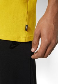 Žluté bavlněné tričko s černou nášivkou Nike na lemování. Černé přiléhavé kalhoty s decentním černým logem Nike po straně.