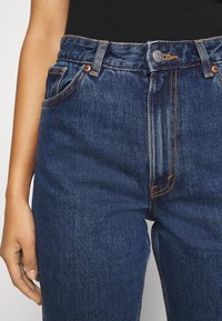 Jeans de mezclilla azul oscuro con cintura alta, cierre de botón en la parte frontal, costuras naranjas y bolsillos laterales. Textura suave con un ajuste clásico.