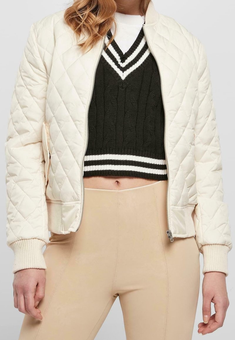 Personne portant un pull court noir et blanc à col en V, un pantalon beige taille haute et une veste matelassée crème avec des poignets côtelés.