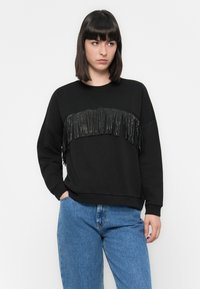 Zwarte sweatshirt met een textuurfranje detail over de borst. Beschikt over lange mouwen en een ronde halslijn, gecombineerd met een blauwe spijkerbroek.