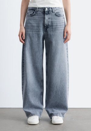 Personne portant un jean en denim bleu clair à coupe ample et des baskets blanches, debout sur un sol gris contre un mur blanc.