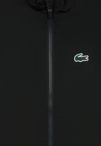 Crna jakna na patent zatvarač od teksturiranog materijala, središnjim patentom i malim zelenim logotipom krokodila na lijevoj strani prsa.